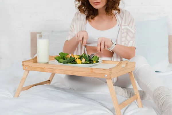 Vista recortada de la mujer embarazada comiendo ensalada con vaso de leche en la bandeja del desayuno en la cama - foto de stock