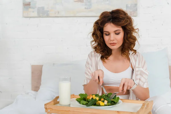 Beautiful woman eating salad near milk on breakfast tray on bed — Stock Photo