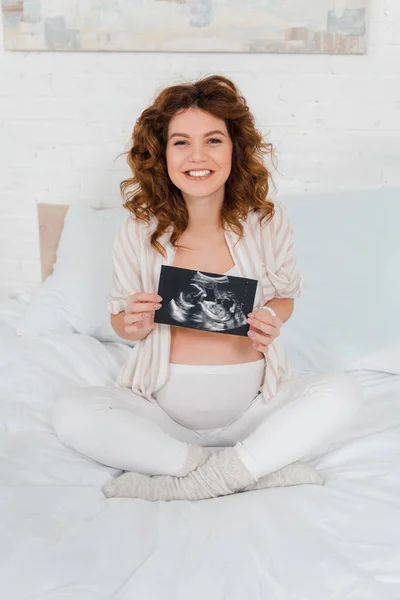 Pregnant woman smiling at camera while holding ultrasound scan of baby on bed — Stock Photo