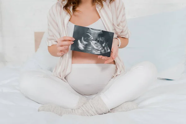 Cropped view of pregnant woman holding ultrasound scan of baby while sitting with crossed legs on bed — Stock Photo