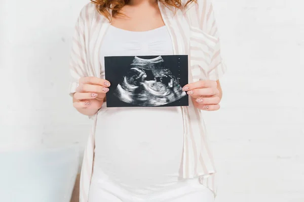 Обрезанный вид беременной девушки, показывающей ультразвуковое сканирование ребенка в спальне — стоковое фото
