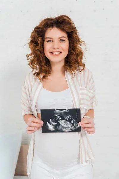 Mujer embarazada sonriendo a la cámara y sosteniendo la ecografía del bebé - foto de stock