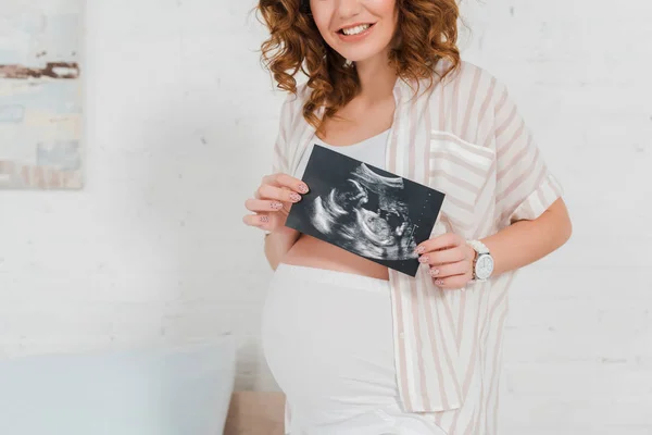 Vista recortada de una mujer embarazada sonriente que muestra una ecografía del bebé en el dormitorio - foto de stock