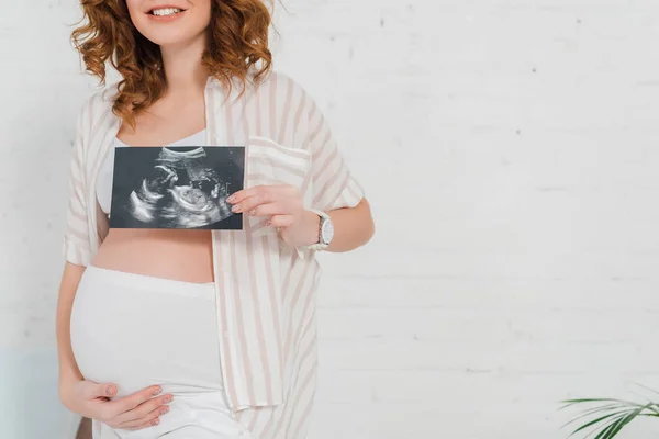 Cropped view of pregnant girl showing ultrasound scan of baby and touching belly — Stock Photo
