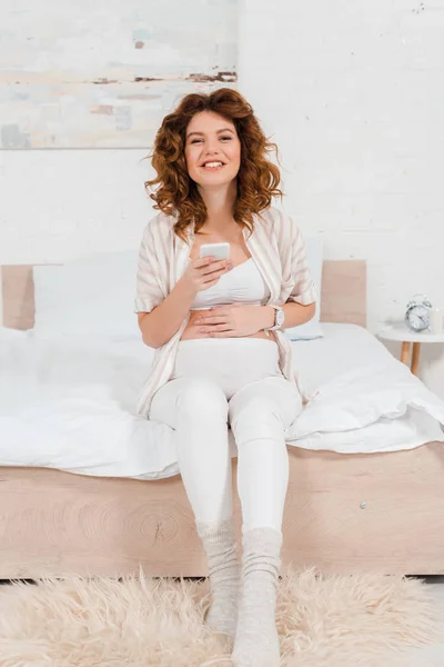 Щаслива вагітна жінка посміхається на камеру і тримає смартфон на ліжку — стокове фото