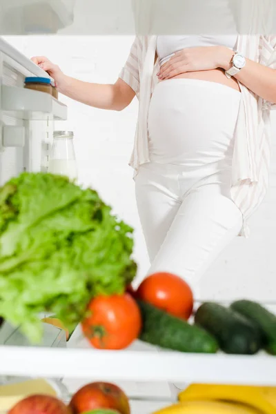 Vista cortada da mulher grávida em pé perto da geladeira aberta com legumes frescos e frutas no fundo branco — Fotografia de Stock