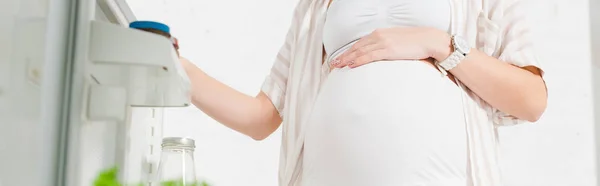 Vista cortada da mulher grávida abrindo porta frigorífico no fundo branco, tiro panorâmico — Fotografia de Stock