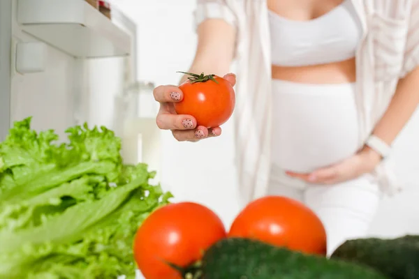 Selective focus of pregnant woman holding tomato near fresh vegetables in open fridge isolated on white — Stock Photo