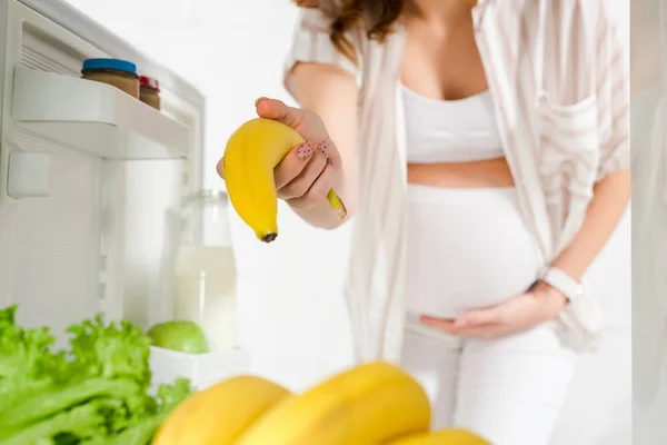 Выборочный фокус беременной женщины, держащей банан в открытом холодильнике на белом фоне — стоковое фото