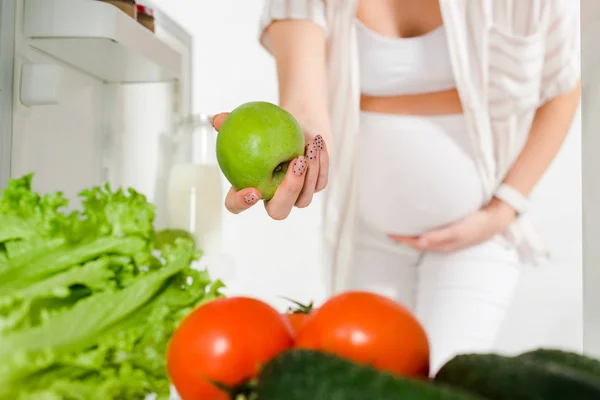 Обрізаний вид вагітної жінки, що тримає яблуко біля свіжих овочів у відкритому холодильнику на білому тлі — стокове фото