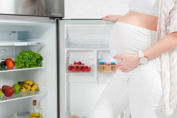 Vista recortada de la mujer embarazada abriendo nevera con verduras frescas y frutas sobre fondo blanco - foto de stock