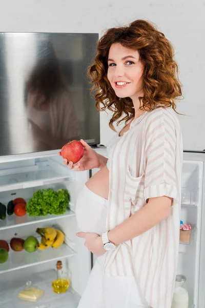 Vista lateral de la mujer embarazada sonriendo a la cámara mientras sostiene la manzana cerca de la nevera abierta con verduras y frutas frescas - foto de stock