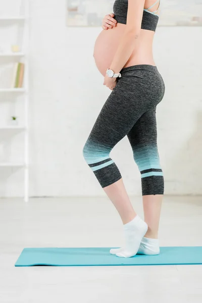 Vista cortada da mulher grávida tocando barriga enquanto estava em pé no tapete de fitness na sala de estar — Fotografia de Stock