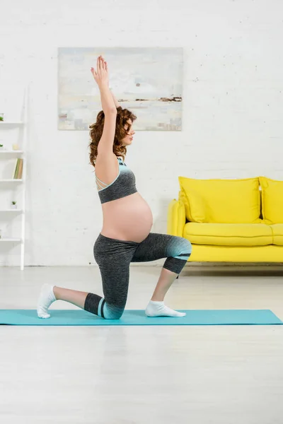 Vista lateral de la chica embarazada practicando yoga asana en la colchoneta de fitness en la sala de estar - foto de stock