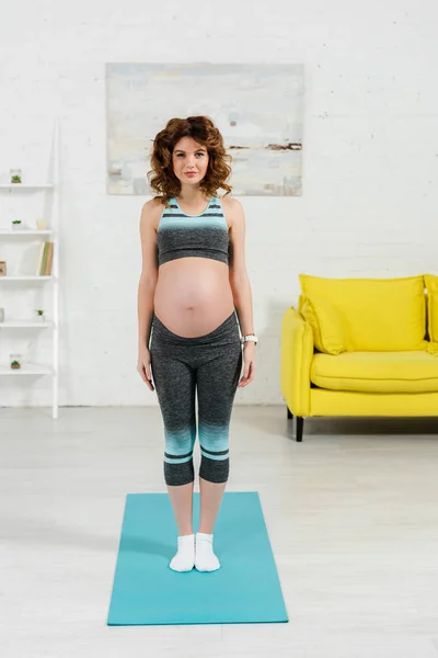 Belle femme enceinte en vêtements de sport regardant la caméra sur tapis de fitness à la maison — Photo de stock