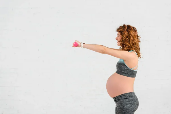 Visão lateral de sorrir mulher grávida treinando com halteres no fundo branco — Fotografia de Stock