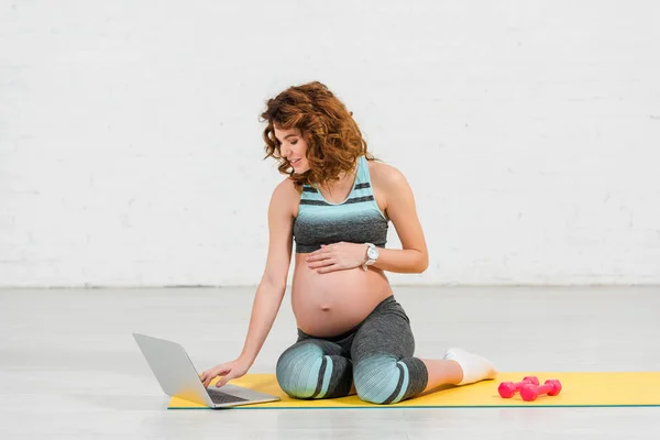 Улыбающаяся беременная женщина с помощью ноутбука на фитнес-коврик рядом гантели на фитнес-коврик дома — стоковое фото