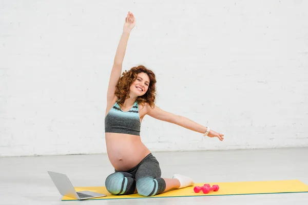 Donna incinta sorridente alla macchina fotografica mentre si allena vicino a laptop e manubri sul tappeto fitness — Foto stock
