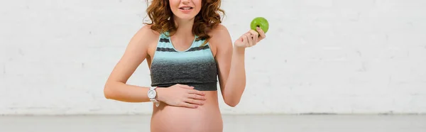 Обрізаний вид усміхненої вагітної жінки, що тримає зелене яблуко, панорамний знімок — стокове фото