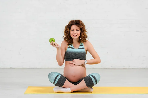 Schwangere hält Apfel in der Hand und lächelt auf Fitnessmatte in die Kamera — Stockfoto