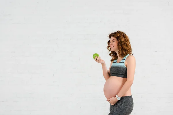 Vista lateral de la mujer embarazada sonriente sosteniendo manzana sobre fondo blanco - foto de stock