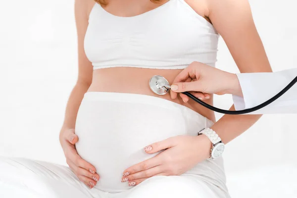 Обрезанный взгляд врача, осматривающего живот беременной женщины стетоскопом, изолированным на белом — стоковое фото