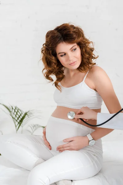 Beautiful pregnant woman sitting on massage table during doctor examining belly with stethoscope in clinic — Stock Photo