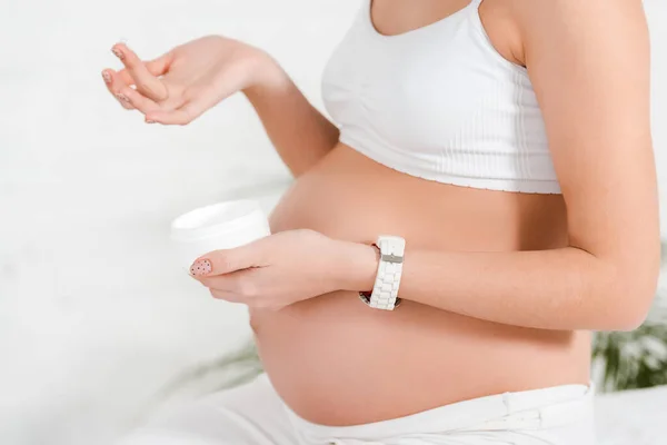 Cropped view of pregnant girl holding jar with cosmetic cream while sitting on bed — Stock Photo