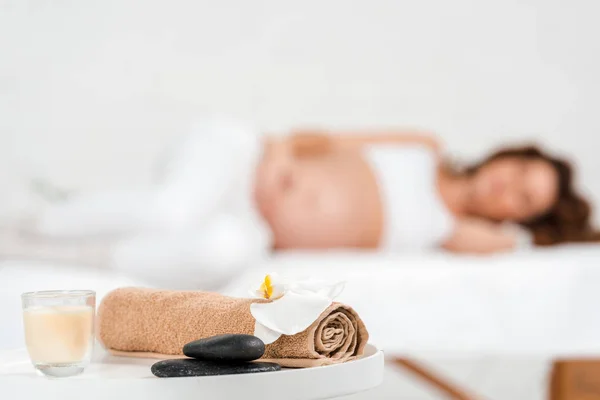 Focus selettivo di pietre, candela con asciugamano e orchidea sul tavolo con donna incinta sul lettino da massaggio nel centro benessere — Foto stock