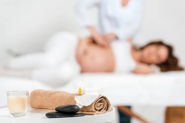 Выборочный фокус камней, свеча с орхидеей на столе и массажист, делающий массаж беременной женщине на массажном столе в спа-центре — стоковое фото