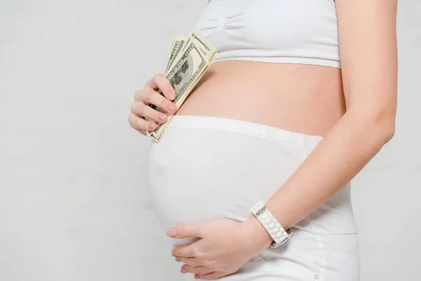 Vista cortada da mulher grávida segurando notas de dólar e tocando barriga em fundo cinza, conceito de maternidade de substituição — Fotografia de Stock