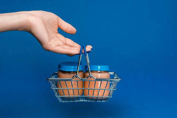 Cropped view of woman holding shopping basket with baby food in jars on blue background — Stock Photo