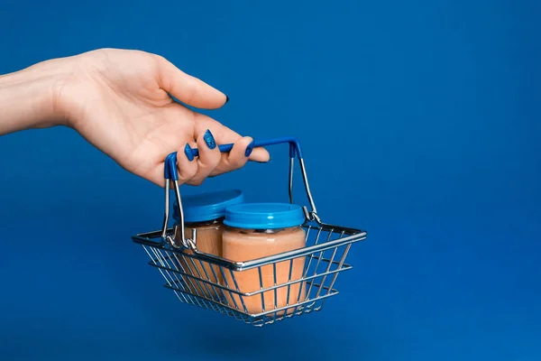 Cropped view of woman holding shopping basket with baby food in jars on blue background — Stock Photo
