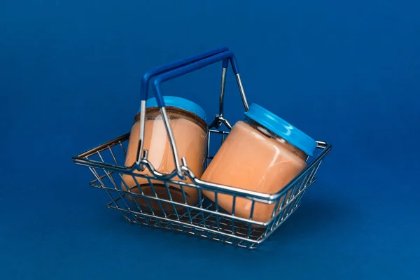 Frascos com comida de bebê na cesta de compras no fundo azul — Fotografia de Stock