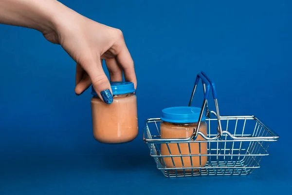 Vue recadrée de la femme mettre dans un bocal panier avec de la nourriture pour bébé sur fond bleu — Photo de stock