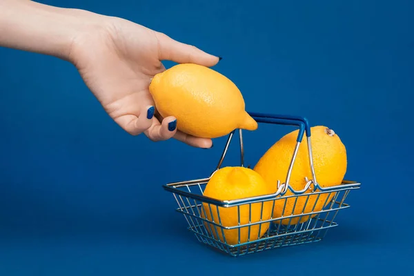 Cropped view of woman putting lemon in shopping basket on blue background — Stock Photo