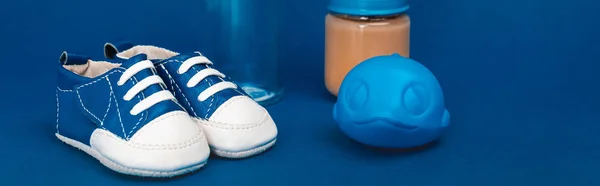 Panoramaaufnahme von Babyflasche, Spielzeug, Babyschuhen, Glas mit Babynahrung vor blauem Hintergrund — Stockfoto