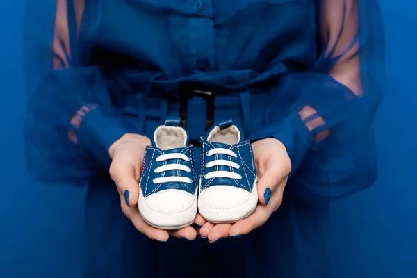 Vista recortada de la mujer sosteniendo zapatos de bebé aislados en azul - foto de stock