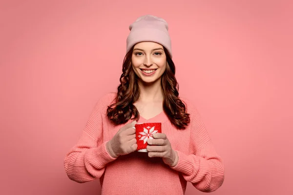 Menina sorridente em chapéu e luvas segurando caneca com bebida aquecida isolado em rosa — Fotografia de Stock