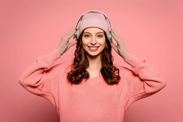 Alegre chica poniendo auriculares inalámbricos mientras sonríe a la cámara en el fondo rosa - foto de stock