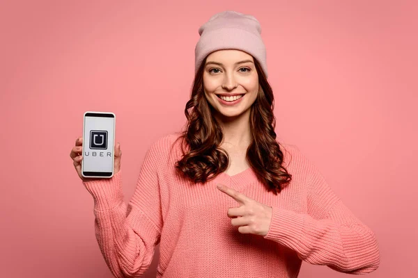 KYIV, UKRAINE - NOVEMBER 29, 2019: smiling girl pointing with finger at smartphone with Uber app on screen isolated on pink — Stock Photo