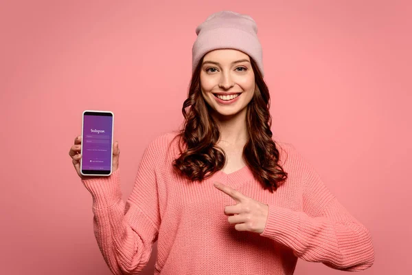KYIV, UCRANIA - 29 de noviembre de 2019: chica sonriente apuntando con el dedo al teléfono inteligente con la aplicación Instagram en la pantalla aislada en rosa — Stock Photo