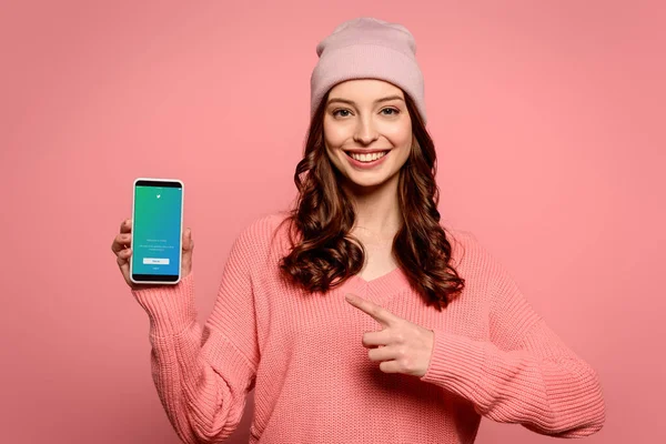 KYIV, UCRANIA - 29 de noviembre de 2019: chica feliz señalando con el dedo al teléfono inteligente con la aplicación de Twitter en la pantalla aislada en rosa - foto de stock