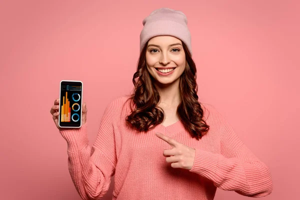 Fröhliches Mädchen zeigt mit dem Finger auf Smartphone mit Diagrammen und Diagrammen auf dem Bildschirm isoliert auf rosa — Stockfoto