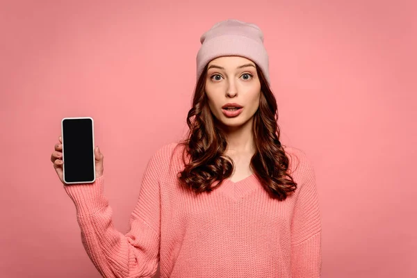 Chica sorprendida mostrando teléfono inteligente con pantalla en blanco mientras mira la cámara aislada en rosa - foto de stock