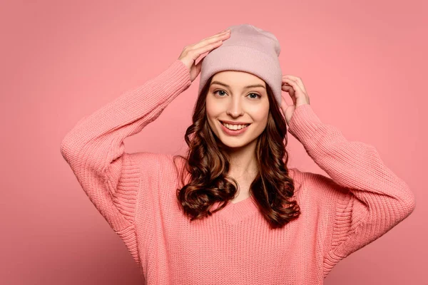 Felice ragazza toccando cappello mentre sorride alla macchina fotografica su sfondo rosa — Foto stock