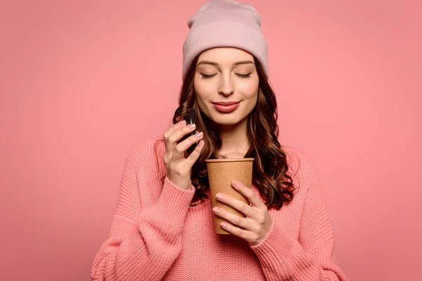 Chica sonriente disfrutando del sabor del café al abrir la taza desechable con los ojos cerrados aislados en rosa - foto de stock