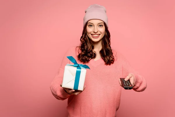 Веселая девушка держит подарочную коробку и телевизор пульт дистанционного управления на розовом фоне — стоковое фото