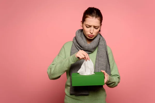 Засмучена, хвора дівчина в теплому шарфі бере паперову серветку з коробки на рожевому фоні — стокове фото