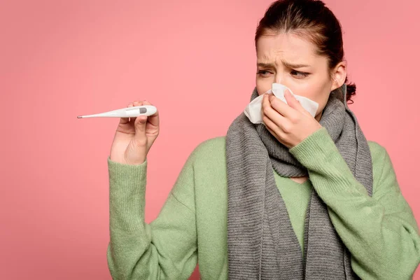 Sick girl in warm scarf wiping nose with paper napkin and showing thermometer isolated on pink — Stock Photo